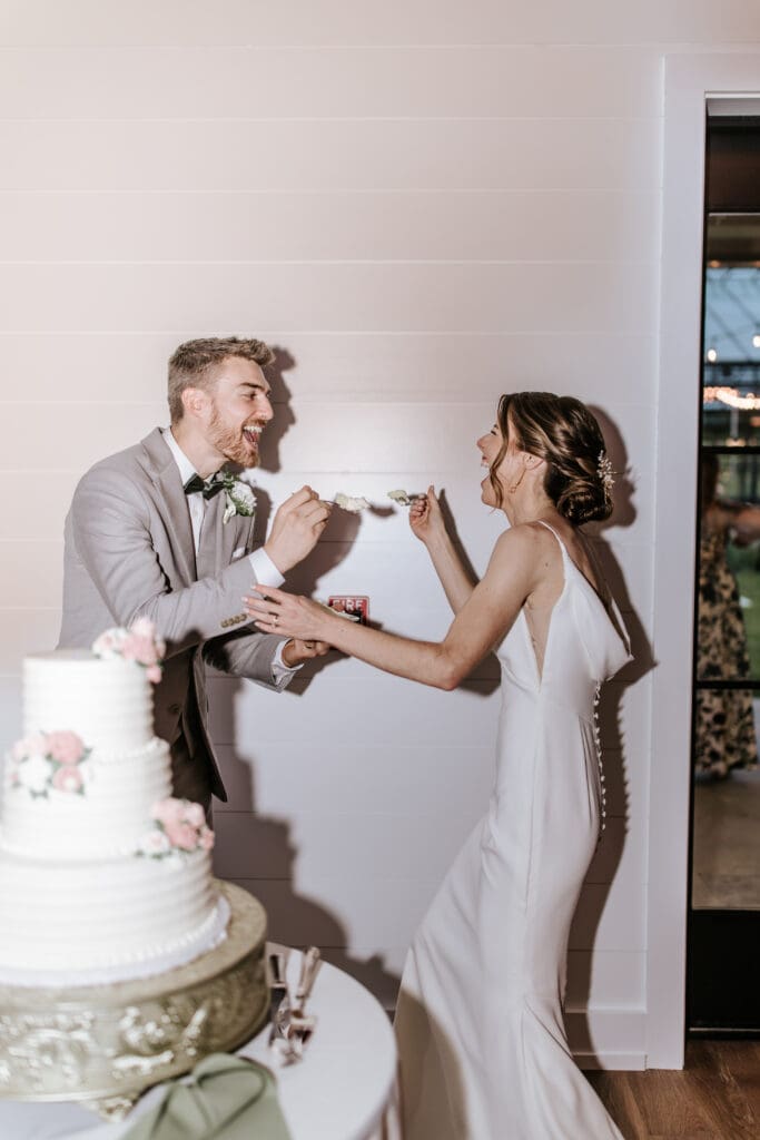 Bride and groom laughing while feeding each other cake during their Westwynd Gardens reception, with a three-tier white cake decorated in pink flowers.