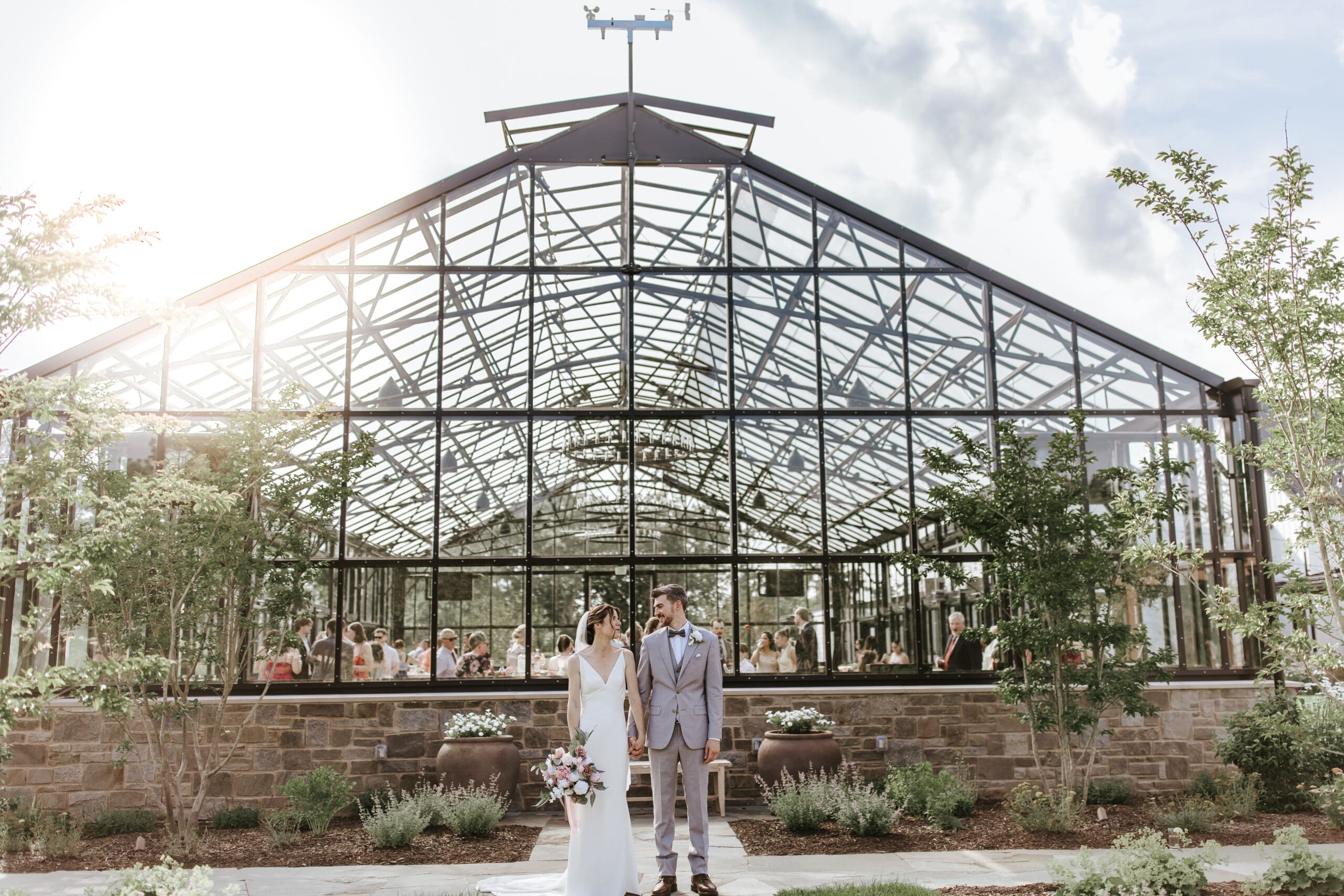 Bride and groom standing in front of the glass greenhouse ceremony space at Westwynd Gardens, a new wedding venue in Honey Brook, PA. The elegant structure reflects the couple and their guests, showcasing the venue’s modern architecture and garden setting.