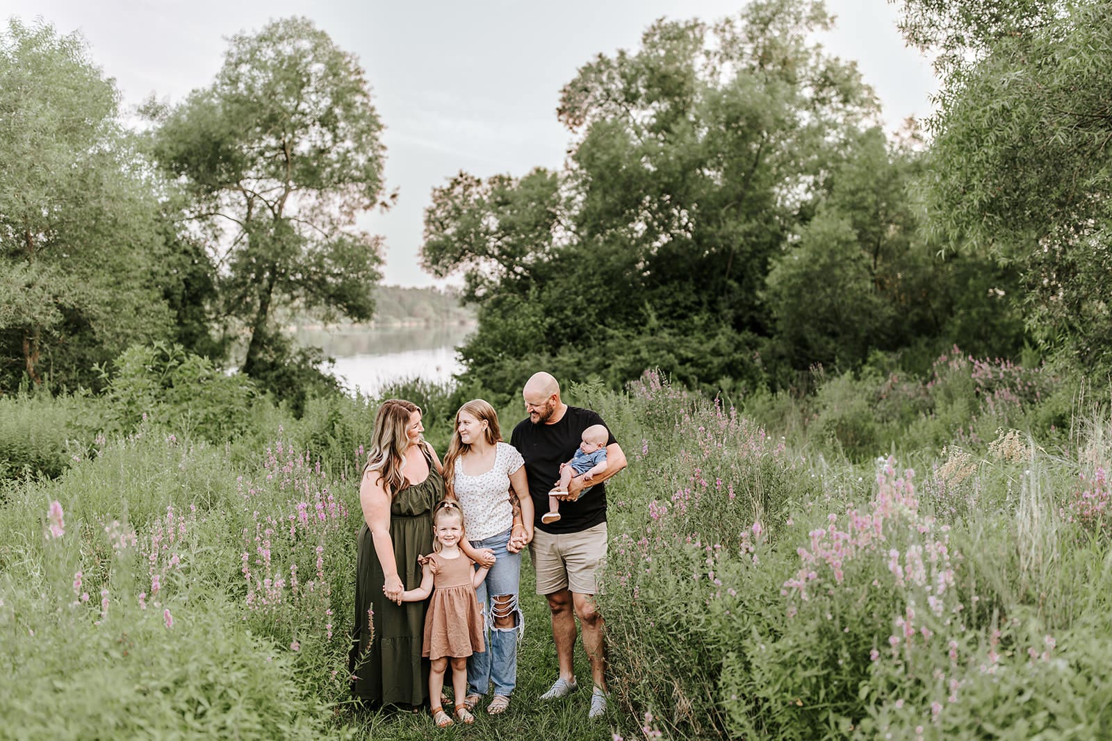West Chester family photographer captures parents with three children, including a newborn, surrounded by wildflowers and a lake view at Struble Lake.
