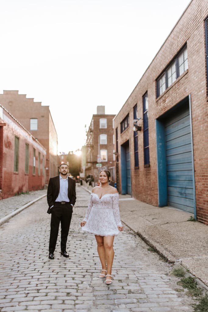 Engaged couple walking on cobblestone street in Old City Philadelphia during their Elfreth’s Alley engagement session, bride leading the way in lace dress.
