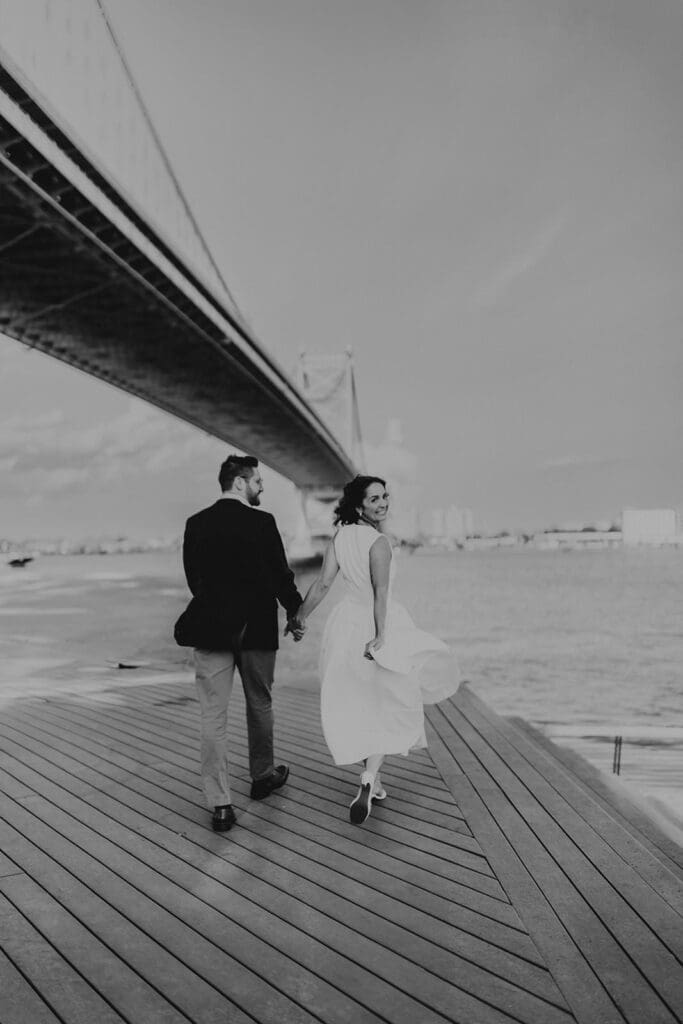 Bride-to-be holding fiancé’s hand while walking along Race Street Pier under the Ben Franklin Bridge during Philadelphia engagement session.