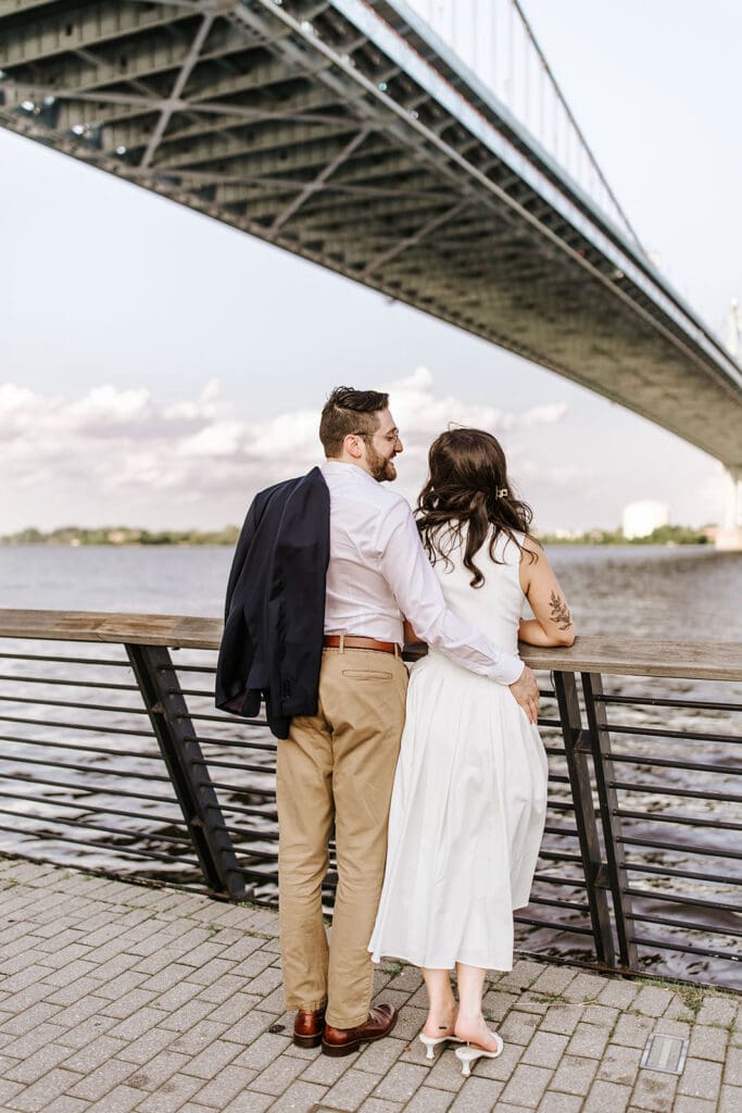 Engaged couple leaning on railing under the Ben Franklin Bridge at Race Street Pier, looking out over the Delaware River in Philadelphia.