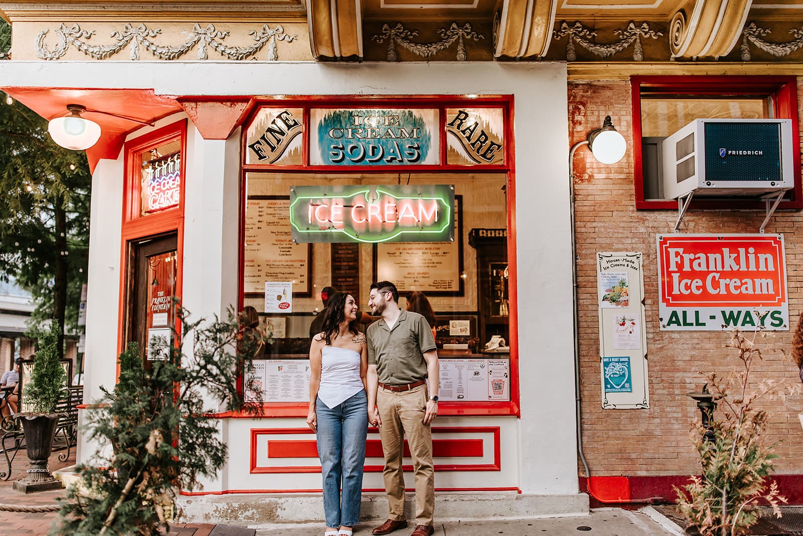 Couple holding hands and smiling outside Franklin Fountain ice cream shop in Old City Philadelphia during their engagement session.