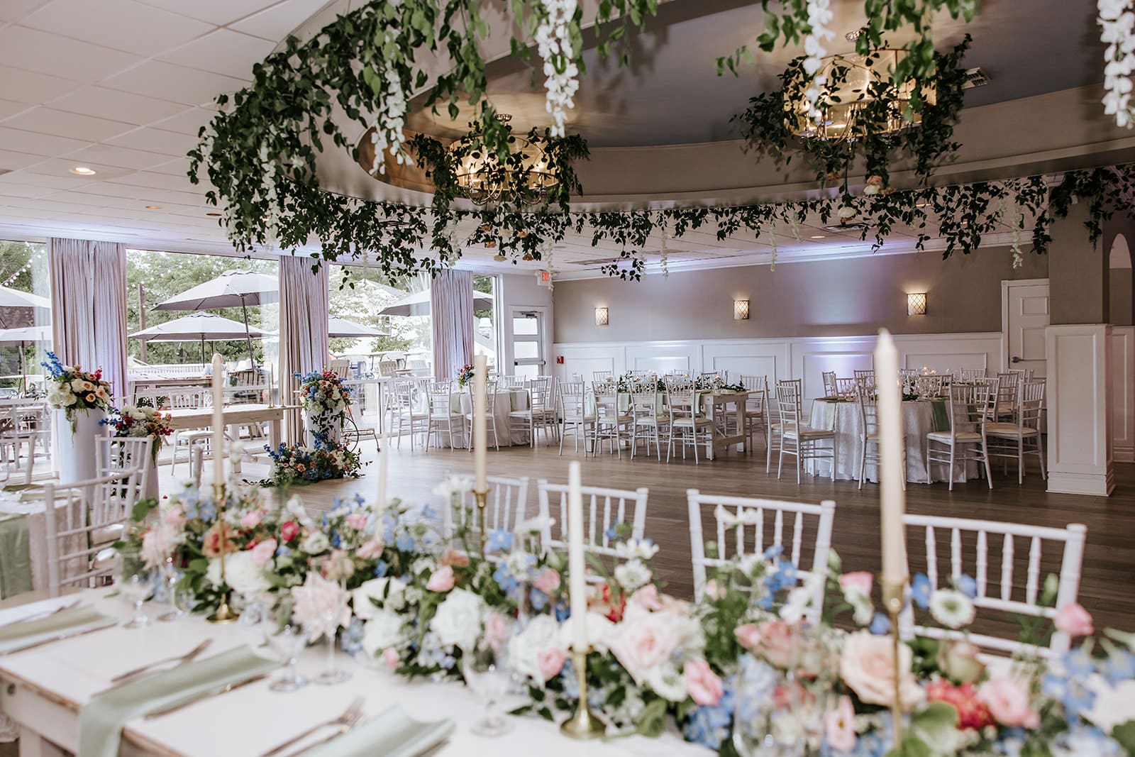 The Grande Ballroom at Ramblewood Country Club decorated for a South Jersey wedding reception with white Chiavari chairs, greenery ceiling installations, and floral centerpieces.