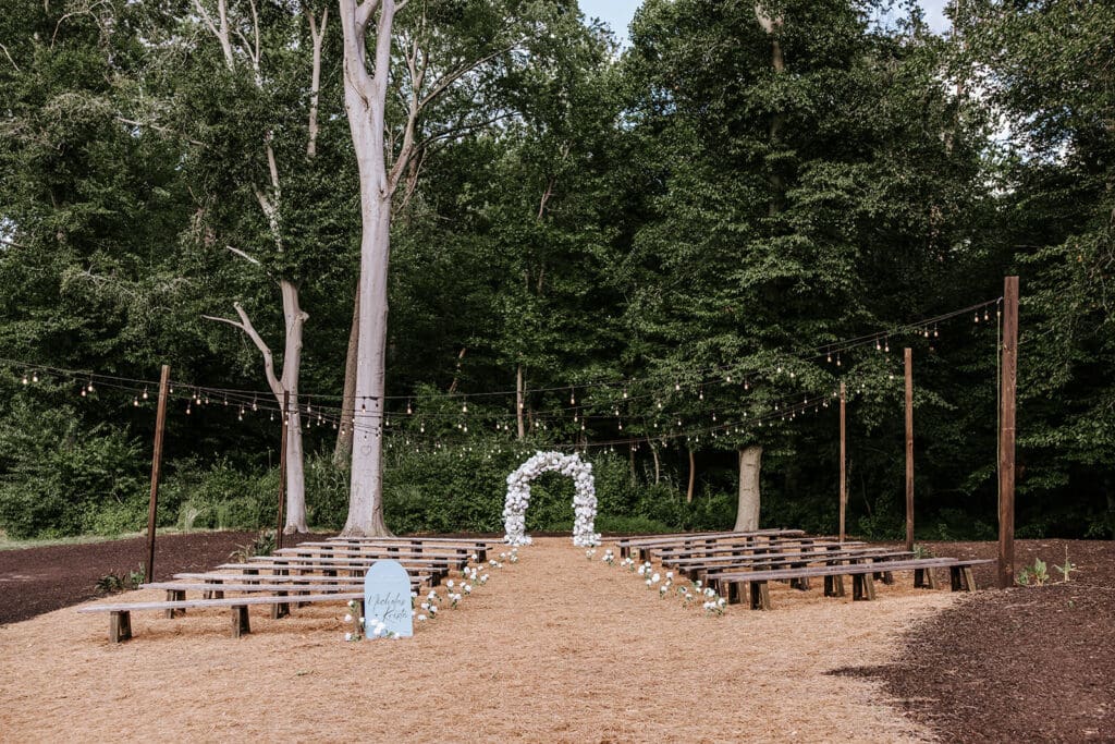 Outdoor wedding ceremony setup at The Oaks at Ramblewood Country Club with string lights, wooden benches, and a floral arch surrounded by tall trees.