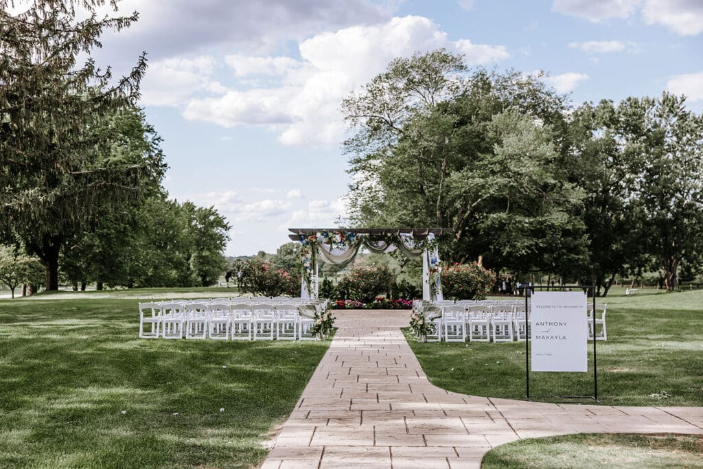 Elegant outdoor ceremony at The Pergola at Ramblewood Country Club with white chairs, floral décor, and golf course views.