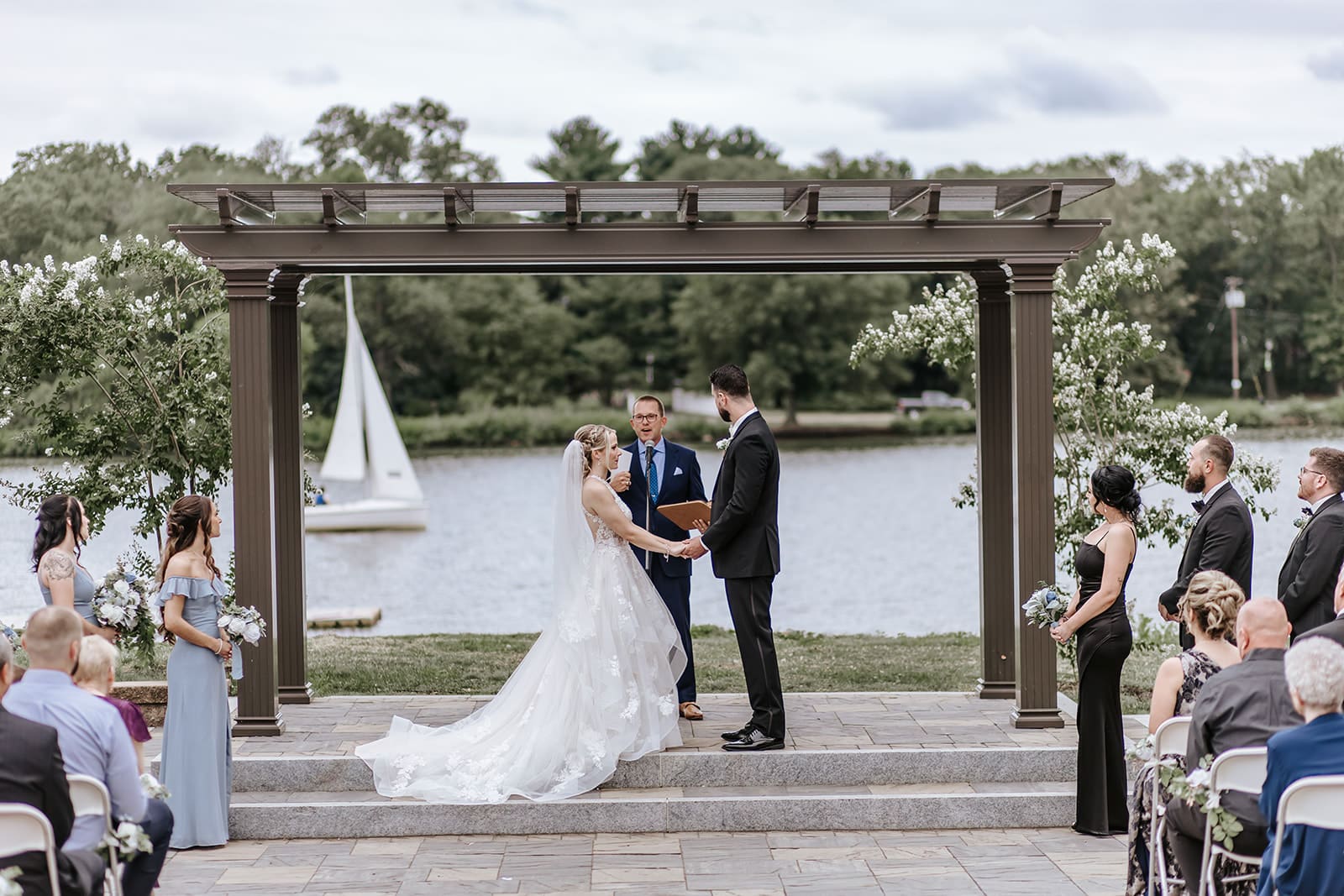 Bride and groom exchanging vows under a pergola during an outdoor ceremony at Camden County Boathouse, with a sailboat passing on the Cooper River in the background.