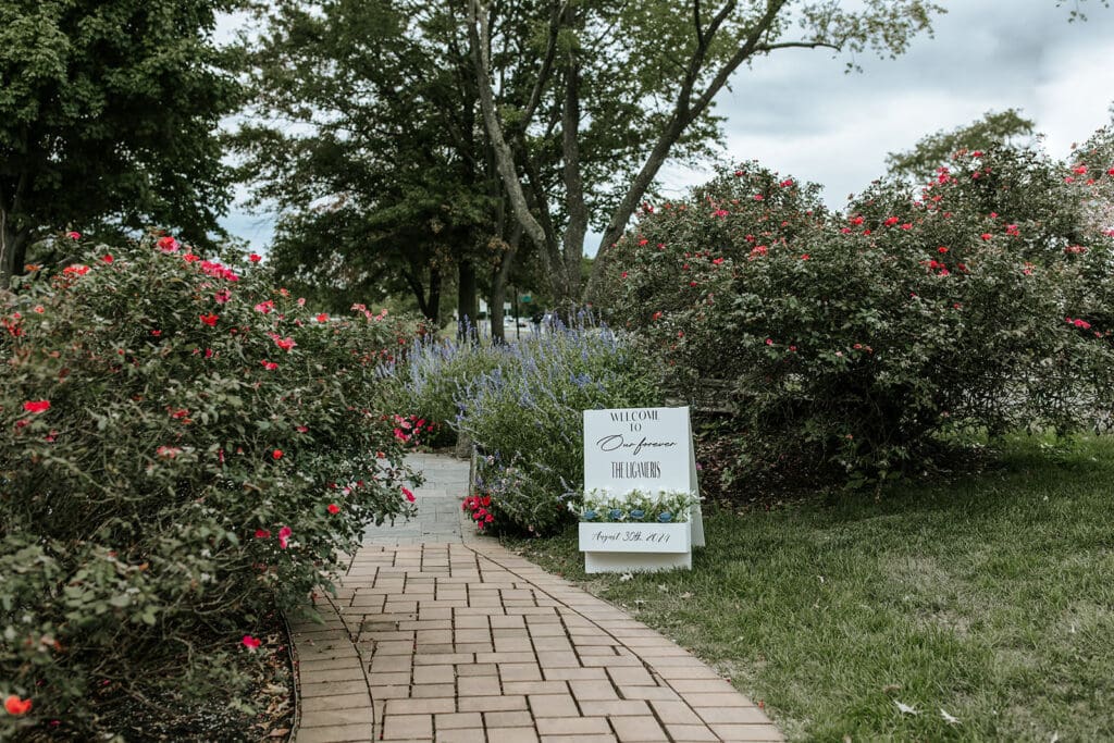 Brick garden path lined with blooming flowers leading to Camden County Boathouse matrimony garden, with a wedding welcome sign displayed.