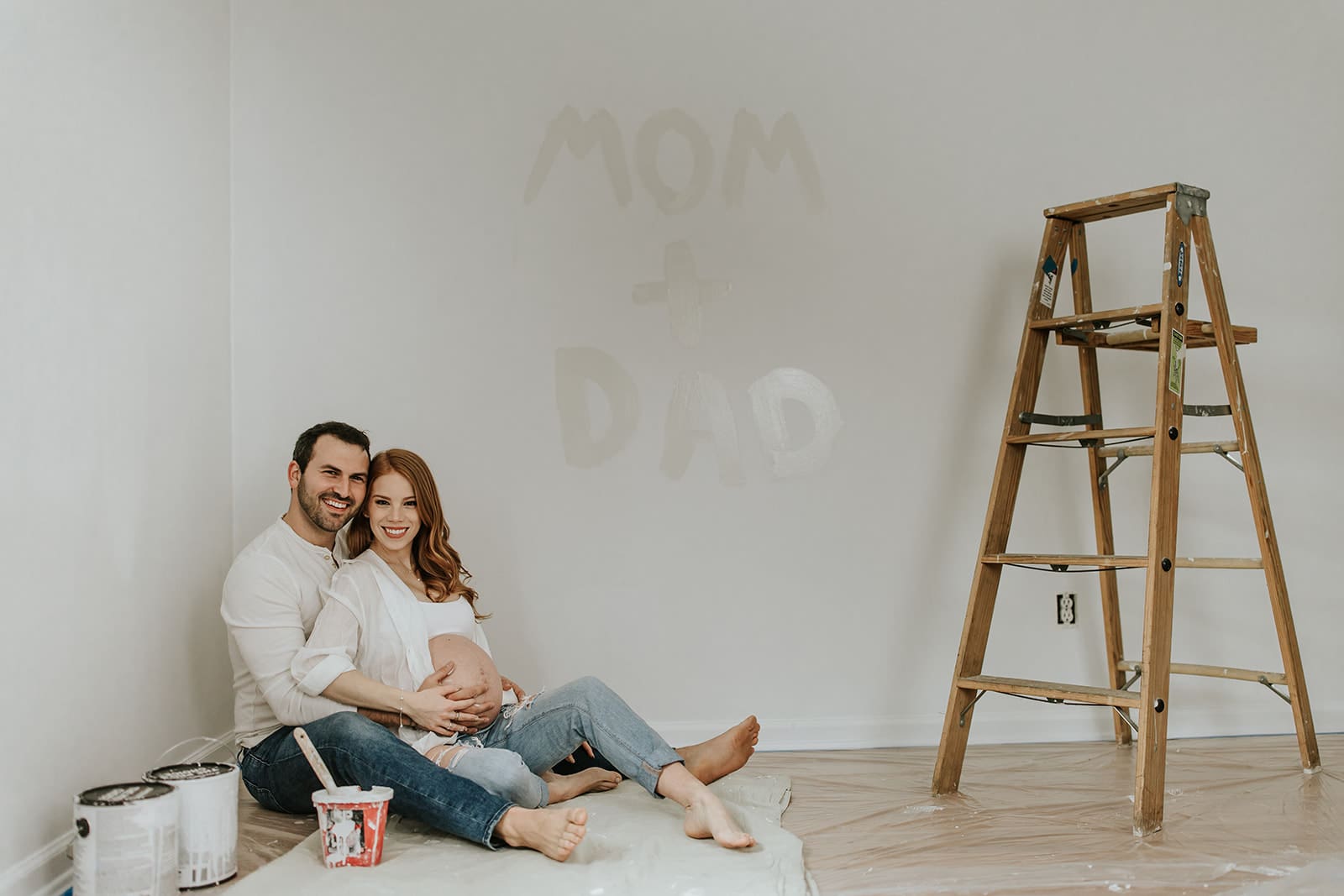 Expecting couple sitting on the nursery floor during their in-home maternity session in West Chester, PA, smiling beside a ladder and paint cans with “Mom + Dad” painted on the wall.