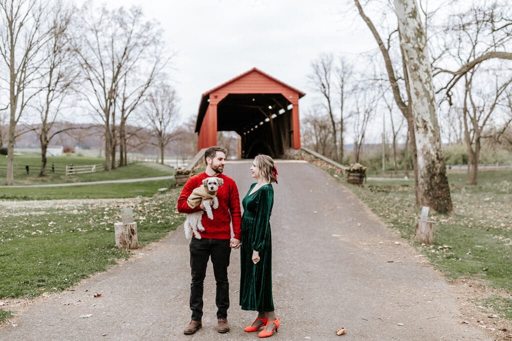 A couple holding hands with their small dog during a Christmas mini session in front of the red covered bridge in Honey Brook, PA; the man wears a red sweater and holds the dog, while the woman in a green dress looks at him and smiles.