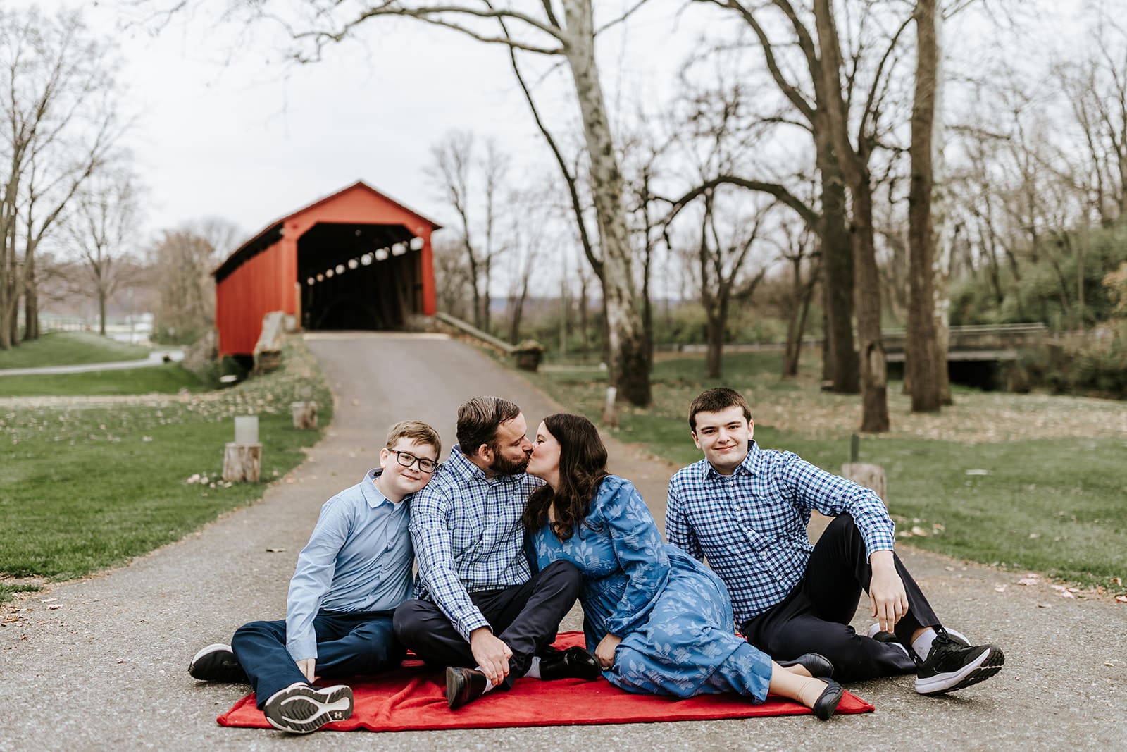 A family sitting on a red blanket in front of the historic red covered bridge in Honey Brook, Pennsylvania; parents kissing while their two sons smile toward the camera during a relaxed outdoor portrait session.