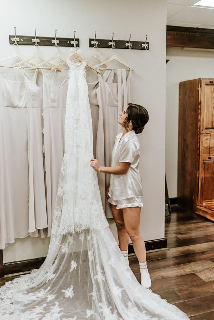 Bride in satin pajamas looking up at her lace wedding dress in the bridal suite at Running Deer Golf Club.