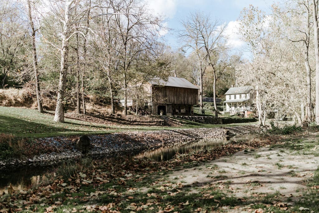 Scenic exterior view of The Mill at Manor Falls in Millersville, Pennsylvania, featuring the historic mill, nearby buildings, and a quiet stream surrounded by autumn trees.