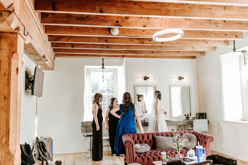 Wedding party getting ready upstairs at The Mill at Manor Falls in Millersville, Pennsylvania, standing beneath exposed wooden beams in the light-filled getting-ready space.