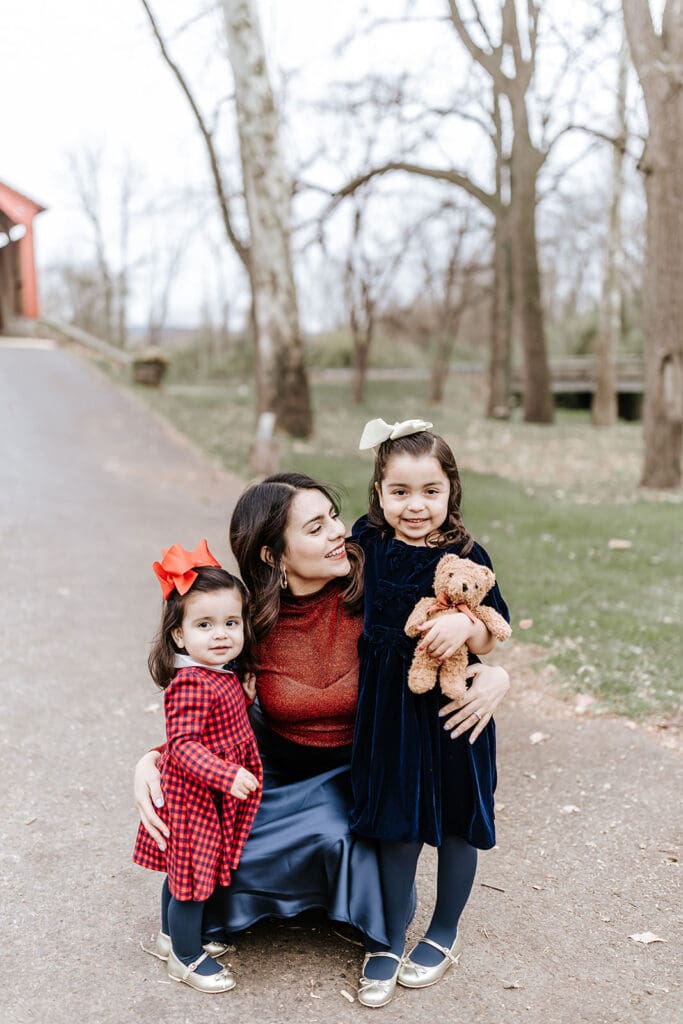 A mother kneeling with her two young daughters during a Christmas mini session in Honey Brook, PA; one daughter kisses her cheek while holding a teddy bear, and the other stands beside her in a red plaid dress with a bow.