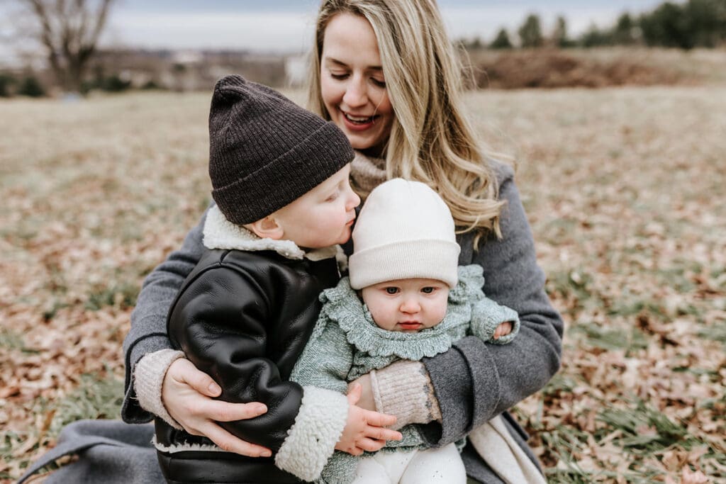 Mother holding her two young children during a winter family photo session at Valley Forge Historic National Park, showcasing natural connection and layered neutral outfits in a relaxed outdoor setting.