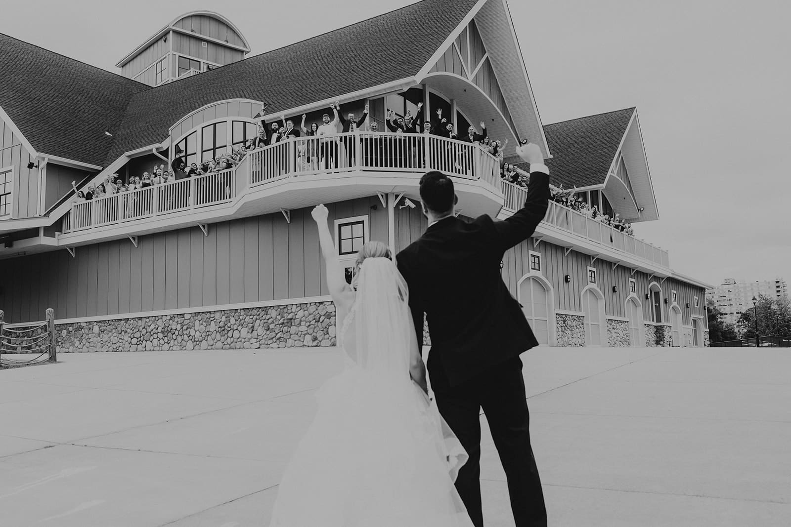 Black and white wedding photo of a newly married couple waving to their guests from outside a barn-style wedding venue after the ceremony.