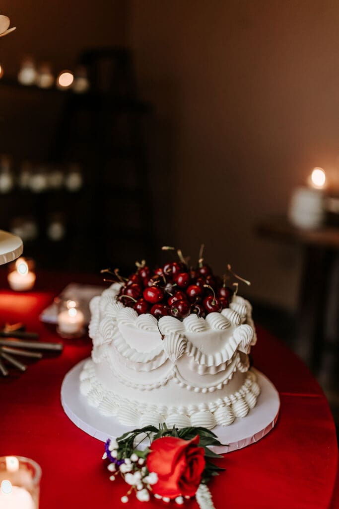 Classic white wedding cake with cherries displayed on a red linen table during the Cherry On Top Wedding Showcase at Running Deer Golf Club in South Jersey.