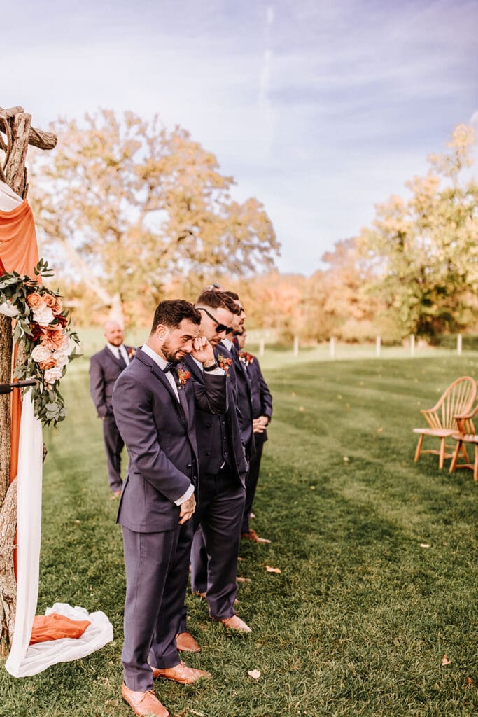 Groom standing at outdoor ceremony with groomsmen waiting for bride, emotional moment before the aisle