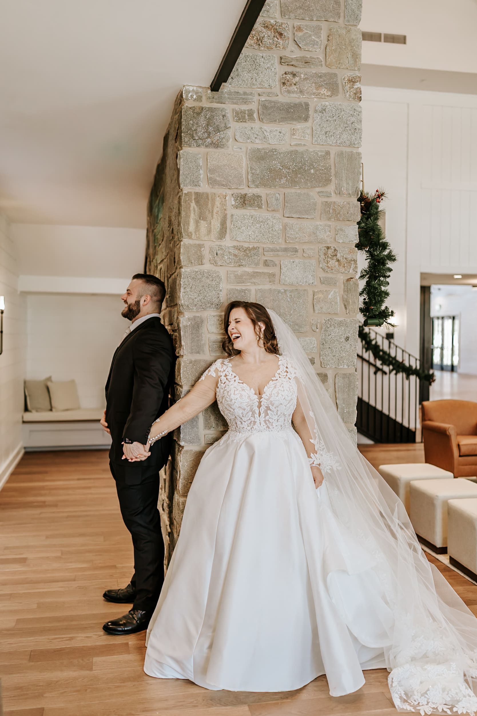 Bride and groom holding hands around a corner during a first look, smiling and laughing before the ceremony