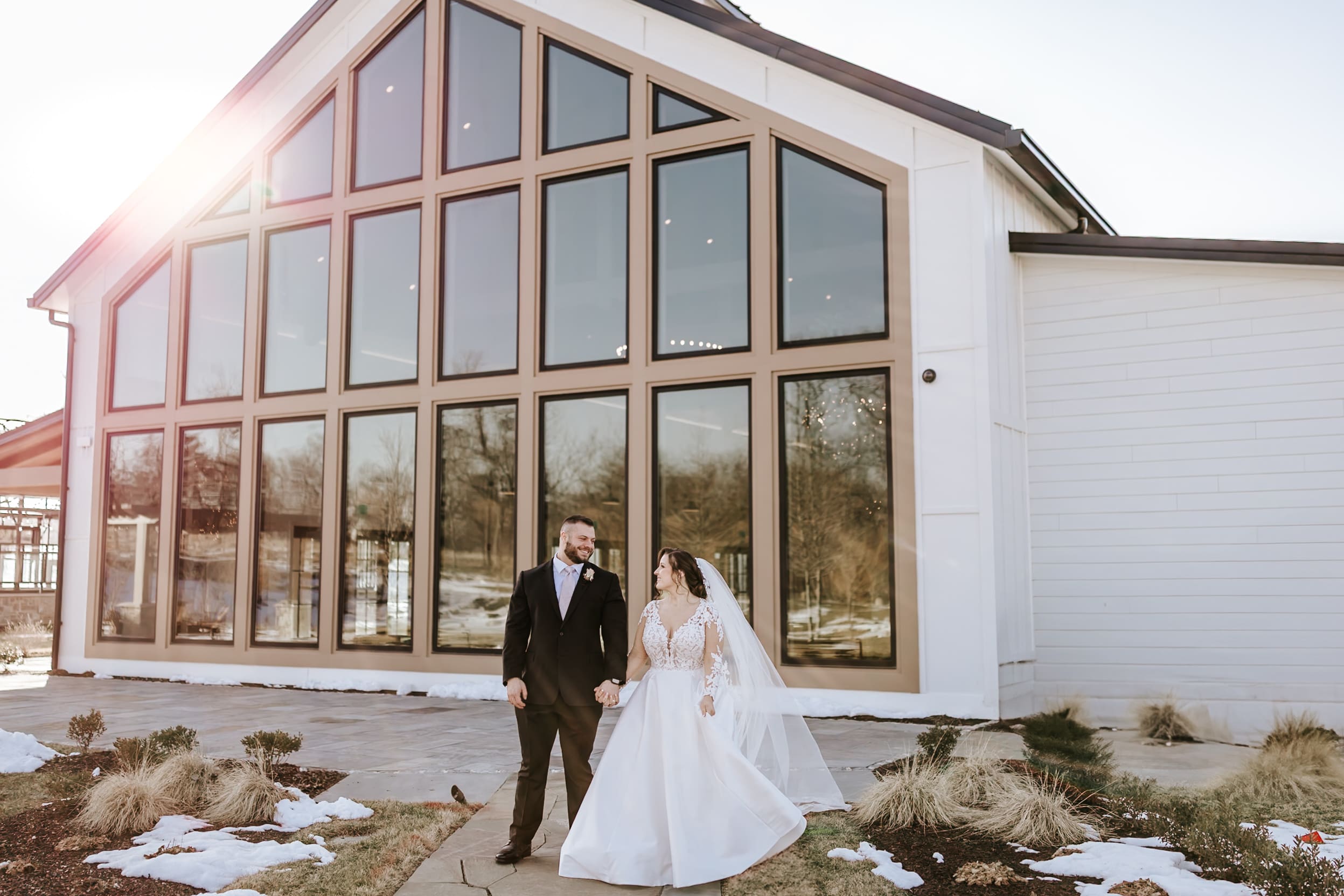 Bride and groom holding hands outside Westwynd Gardens in Honey Brook, PA during a winter wedding, standing in front of the modern greenhouse with large windows and patches of snow on the ground.