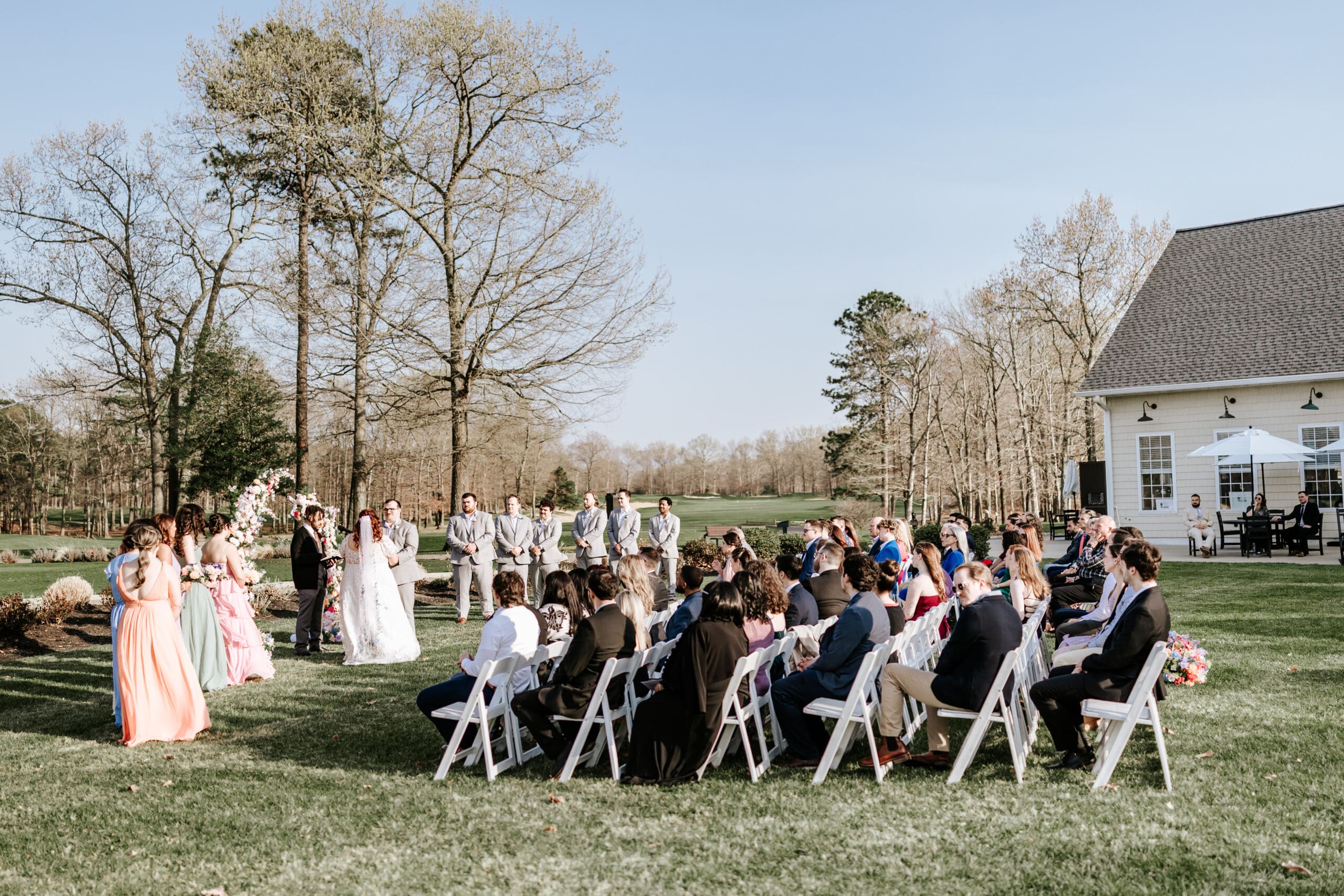 Outdoor wedding ceremony on the grass at Running Deer Golf Club in South Jersey, with pastel bridesmaid dresses, floral arch, and golf course views in the background on a sunny spring day