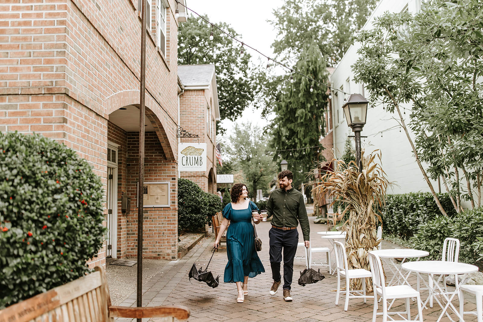 Engaged couple strolling through downtown Haddonfield carrying umbrellas and coffee after a rain shower.