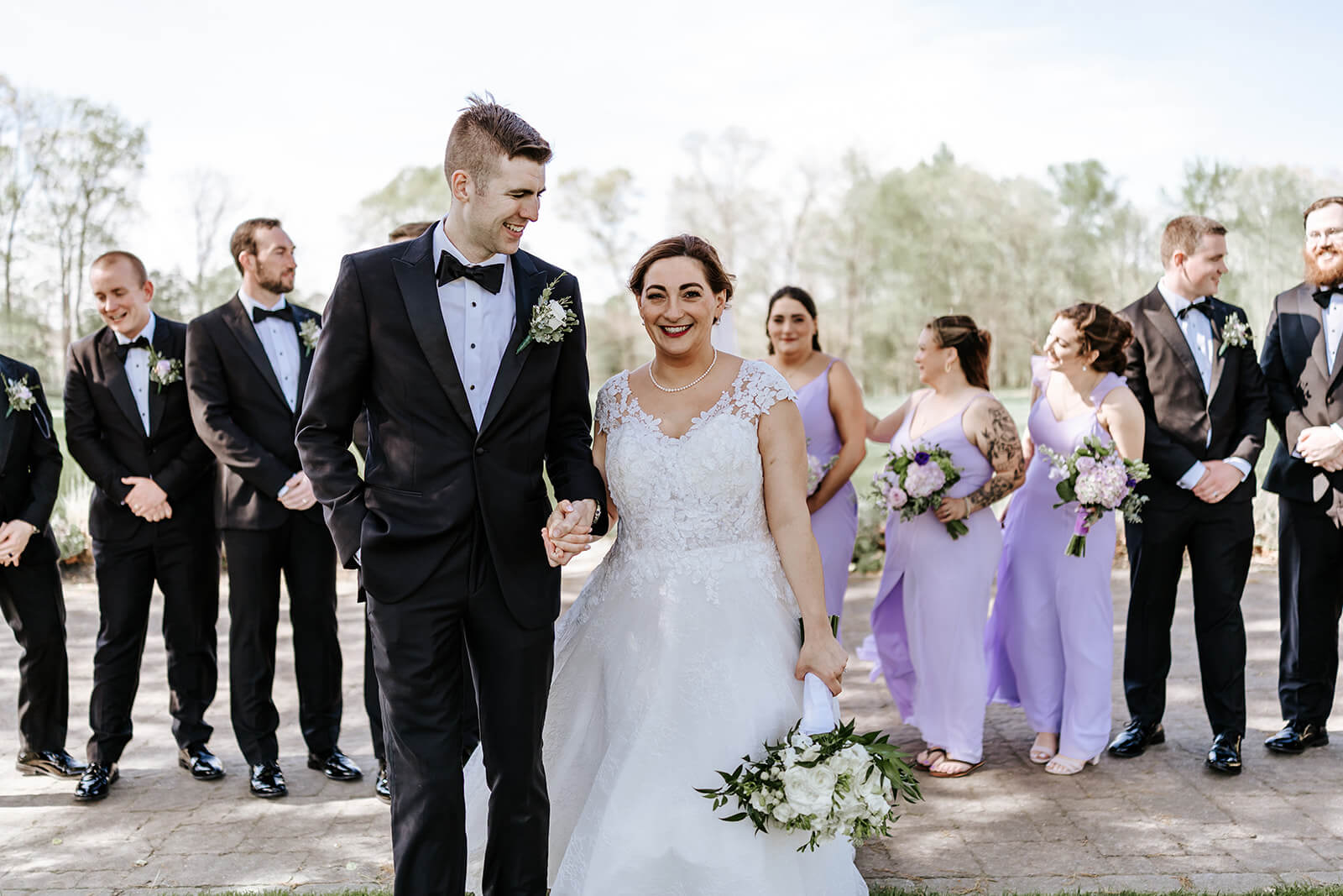 Wedding party portrait with bride, groom, bridesmaids in lavender dresses, and groomsmen in black tuxedos.