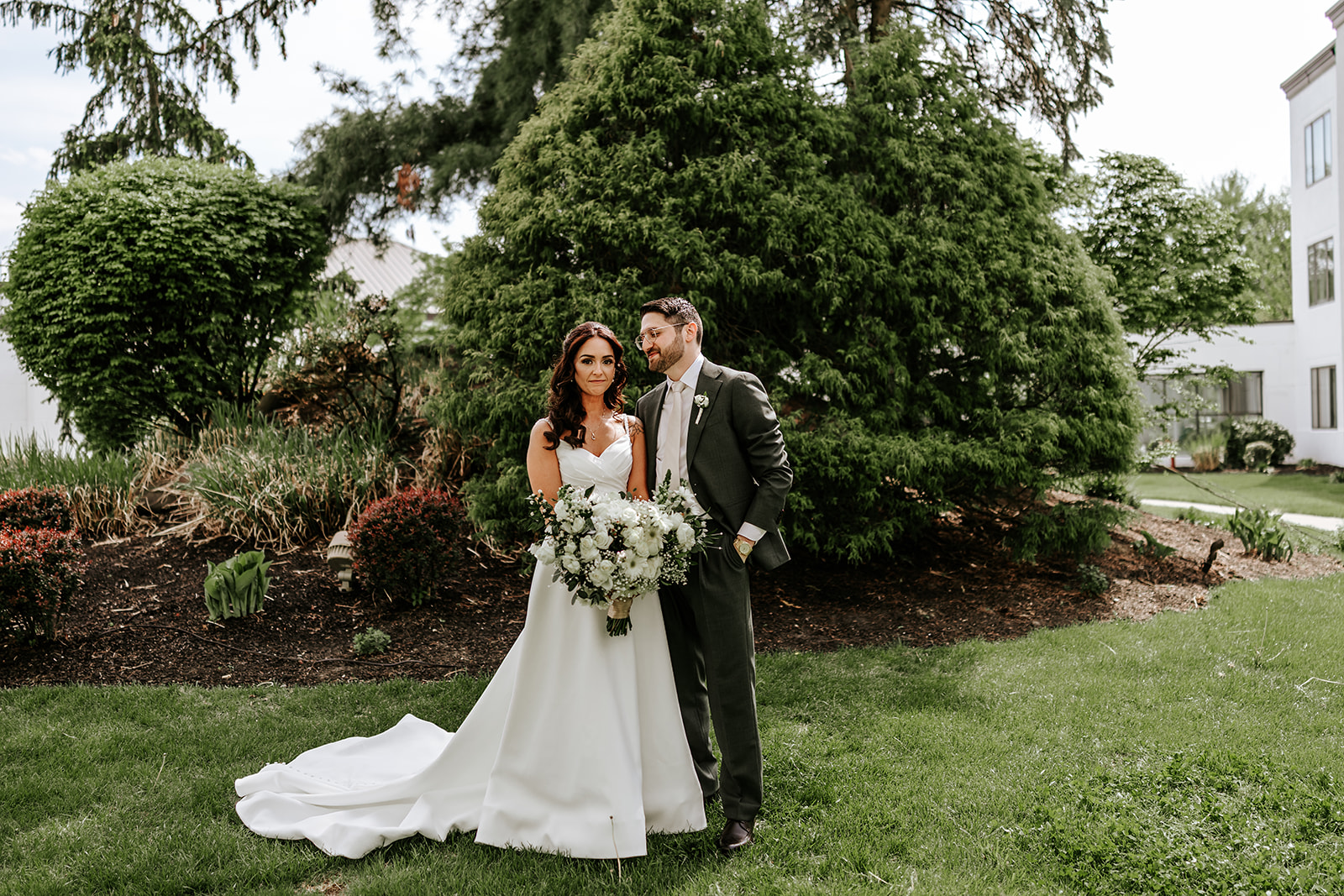 Full-length portrait of the bride and groom standing together outdoors with the bride's wedding dress train and bouquet.