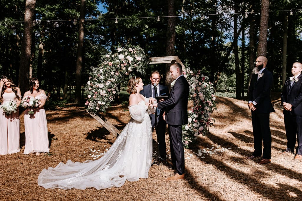 bride wearing whimsical wedding dress smiling while putting ring on groom's finger in front of a hexagon arch with floral arrangement, surrounded by bridal party